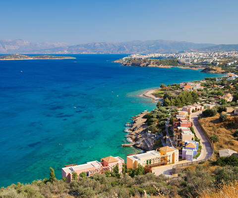 Panoramic coastal view with colorful buildings, blue waters, and distant mountains in Crete